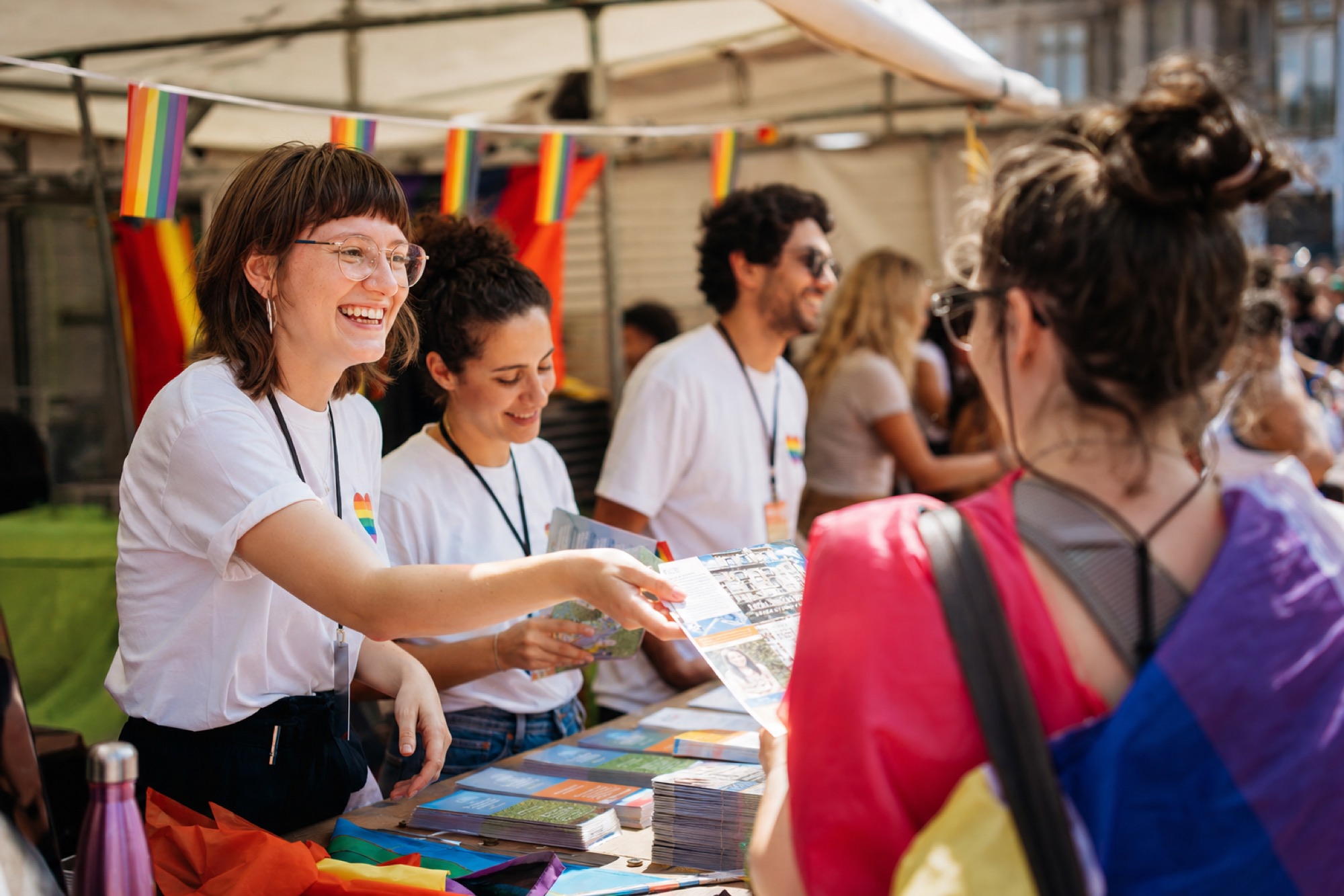 Volunteers at a community booth handing out printed guides during a Pride street festival